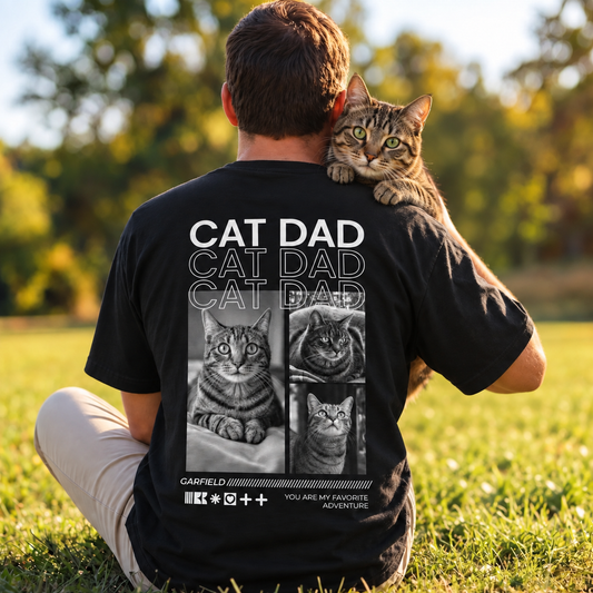 Camiseta negra con el texto "Cat Dad" y motivos de gatos, llevada por una persona con un gato.