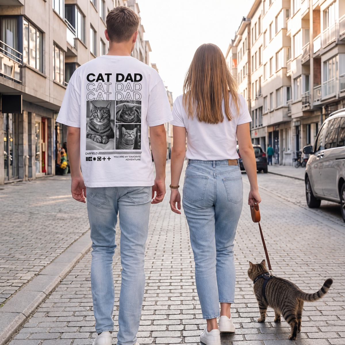 Camiseta blanca con estampado de gatos en la espalda, dos personas y un gato paseando por una calle.