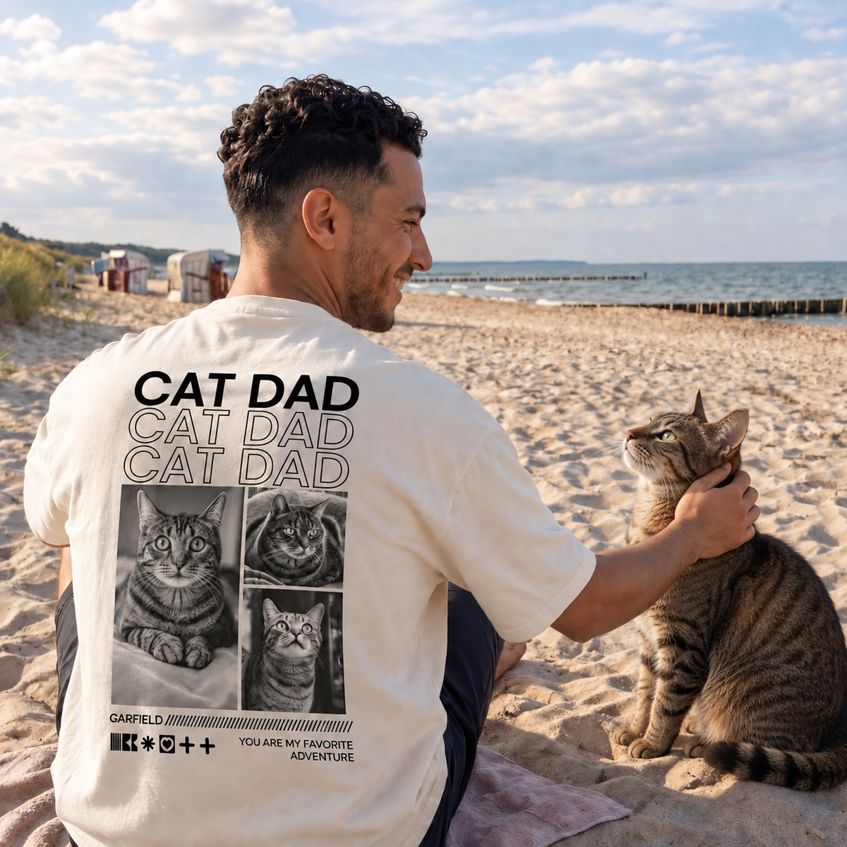 Camiseta beige con el texto "Cat Dad" y motivos de gatos en la espalda, llevada por un hombre en la playa.