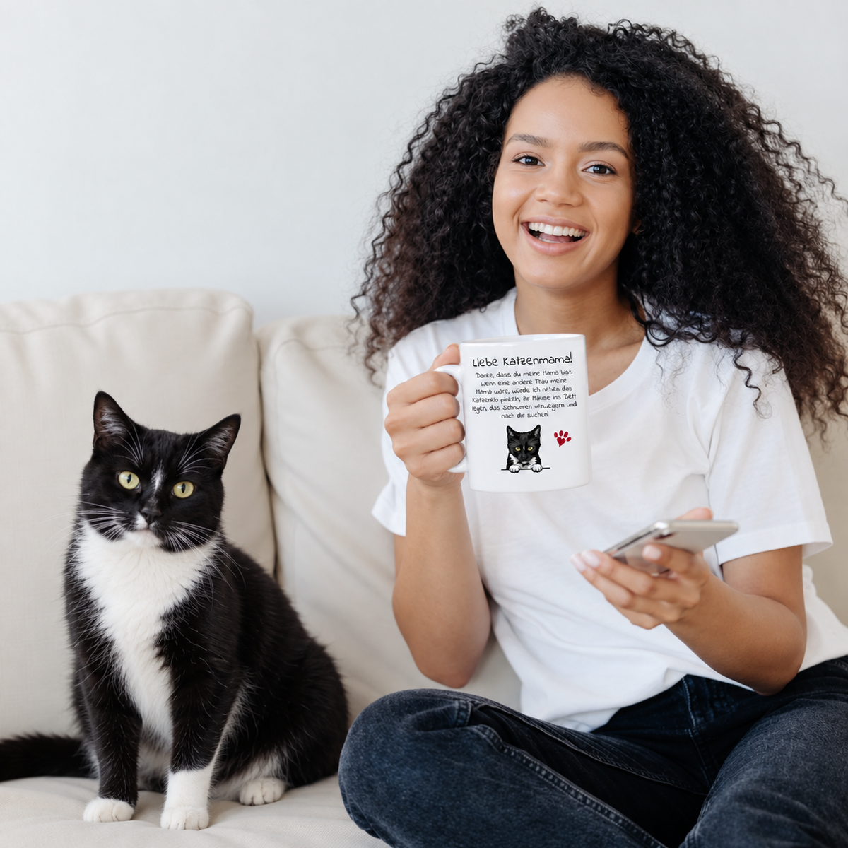 Joven con pelo rizado sosteniendo una taza personalizada con motivo de gato, un gato negro a su lado en el sofá.