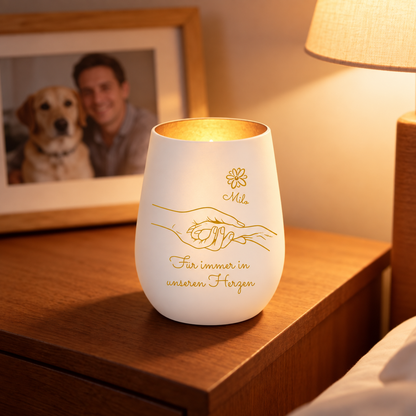 Vela conmemorativa de vidrio personalizada con letras doradas y diseño de manos, brilla suavemente en una mesilla.