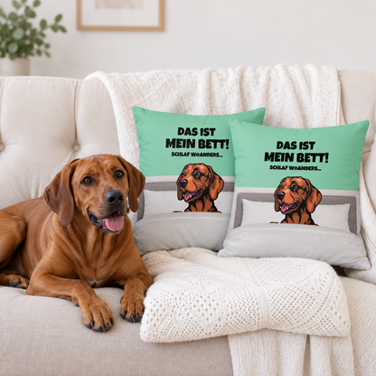 Cojín azul claro y gris con motivo de perro y el texto "Das ist mein Bett!" en un sofá. Perro tumbado al lado.