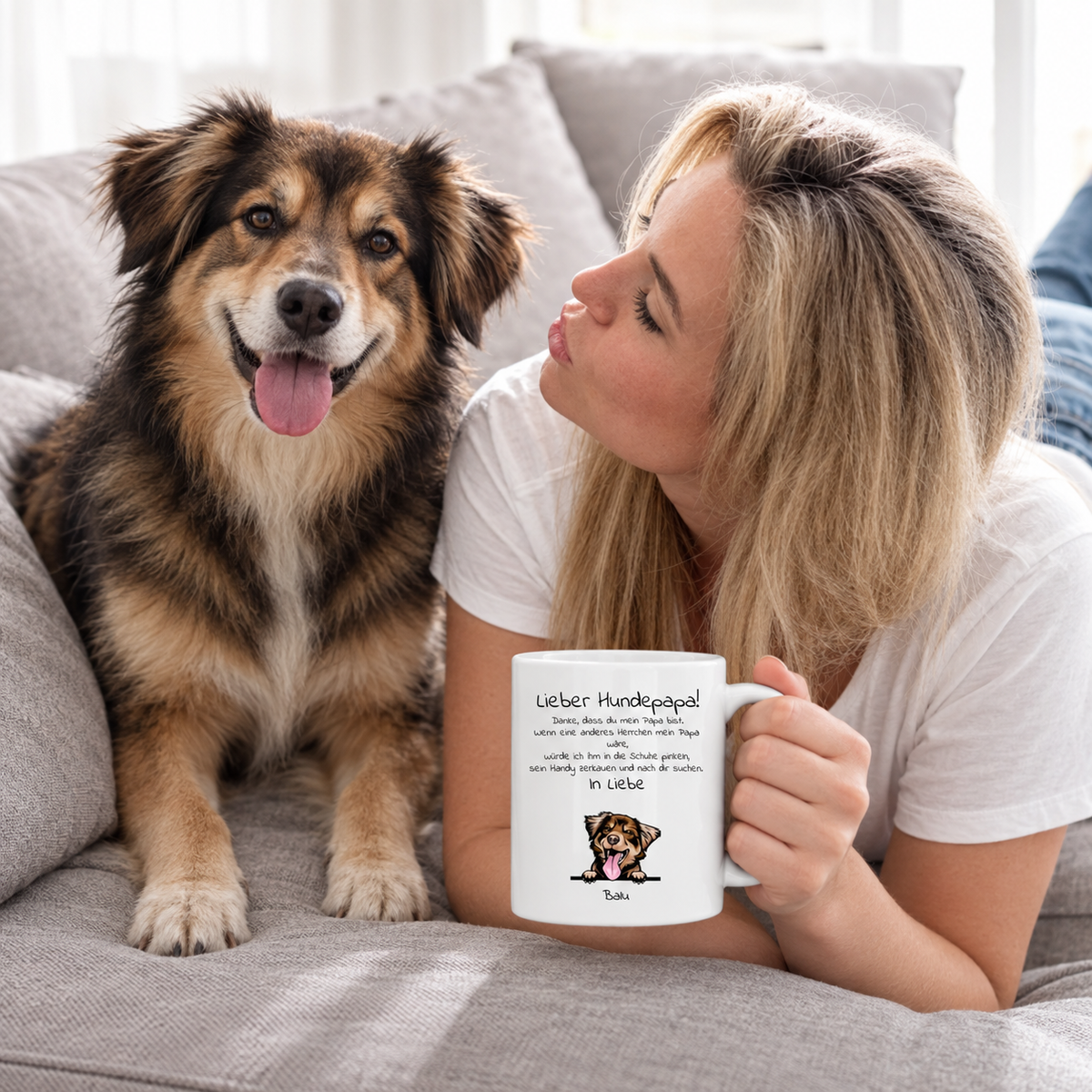 Mujer con taza personalizada "Lieber Hundepapa" y perro en un sofá gris. Taza blanca con texto e ilustración.
