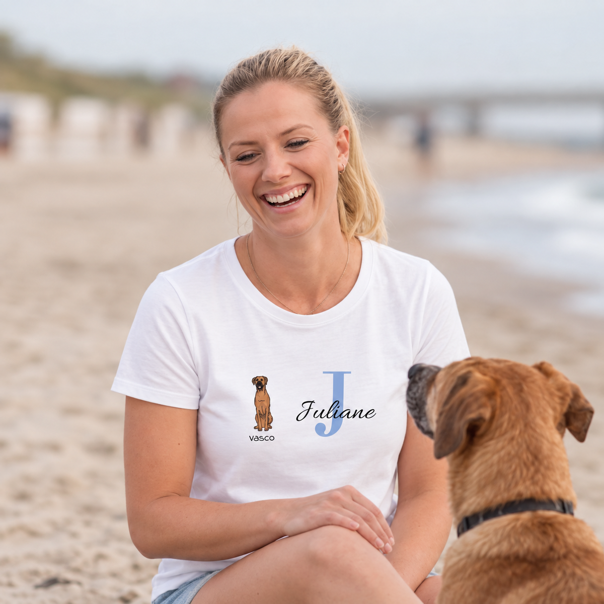Camiseta blanca con gráfico de perro y el nombre Juliane en letras azules. Mujer sentada en la playa.