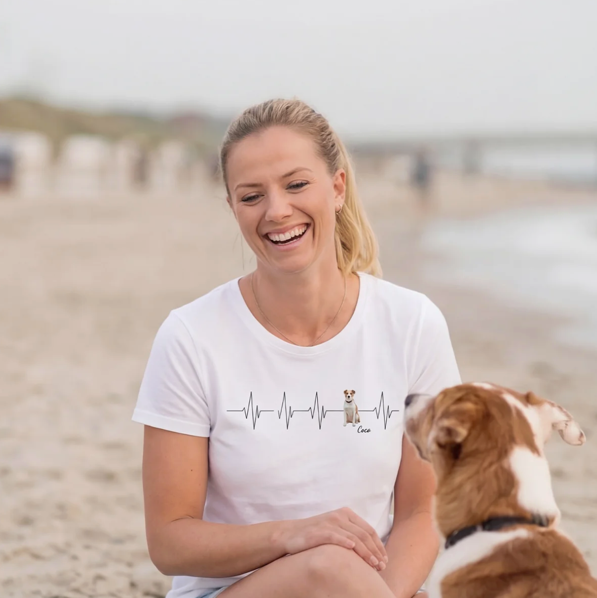 Camiseta blanca con patrón de latido y motivo de perro, mujer sentada en la playa.