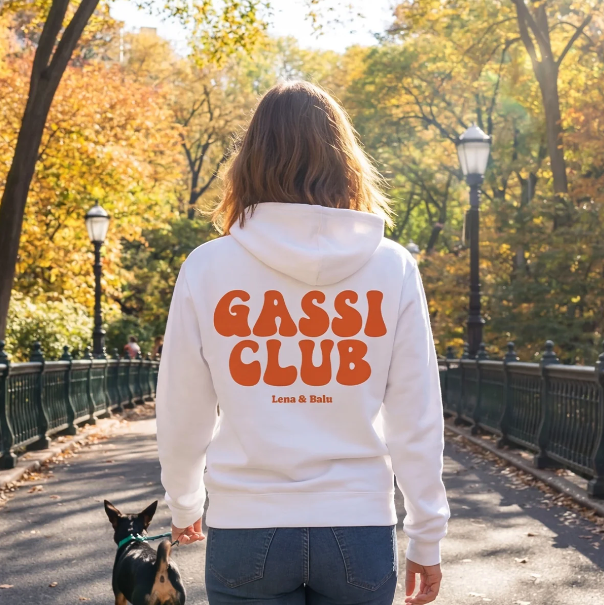 Sudadera con capucha blanca con el texto "Gassi Club" y "Lena & Balu" en la espalda. Persona paseando un perro en el parque.