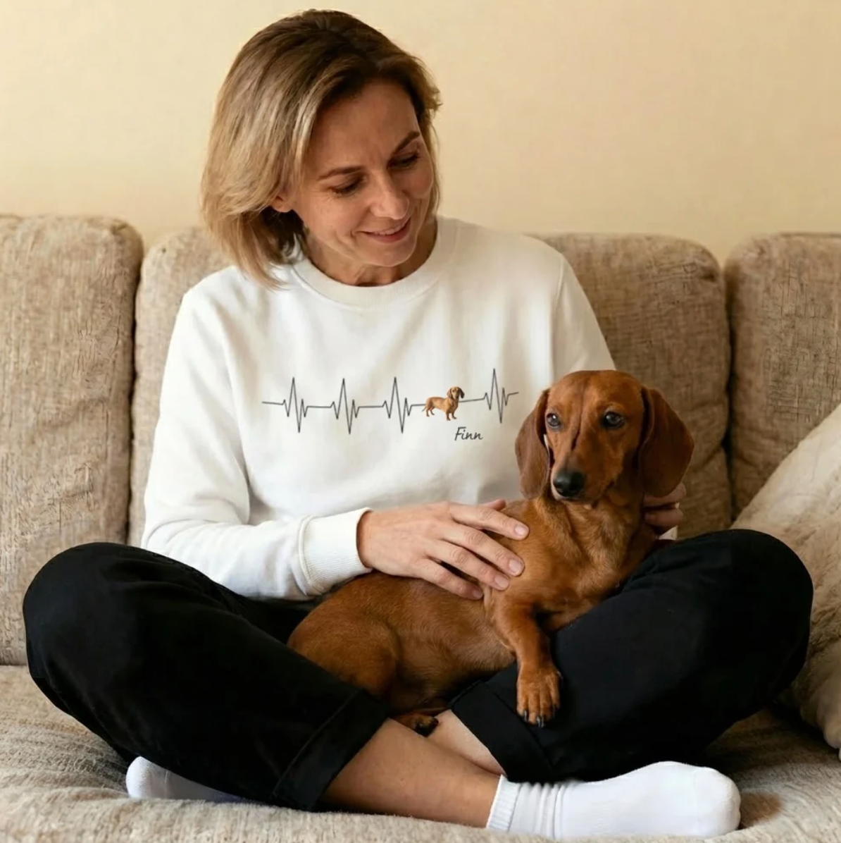 Sudadera blanca con motivo de latido y un perro, mujer sonriente sentada en un sofá con un teckel.