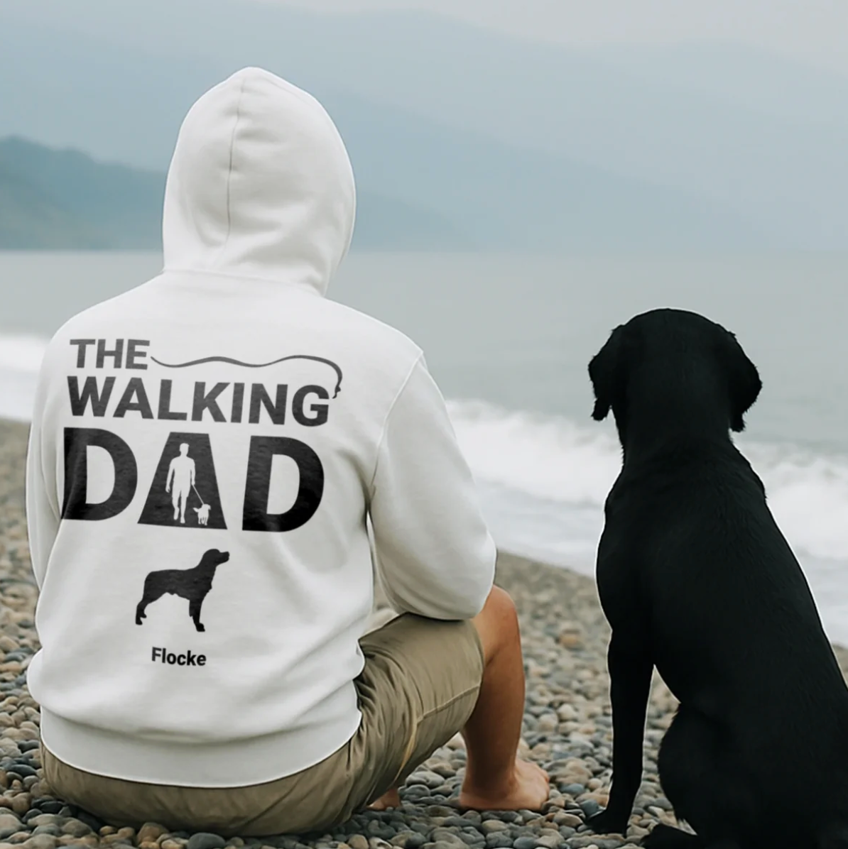 Sudadera con capucha blanca con "THE WALKING DAD" en la espalda y motivo de perro, llevada por un hombre sentado en la playa.