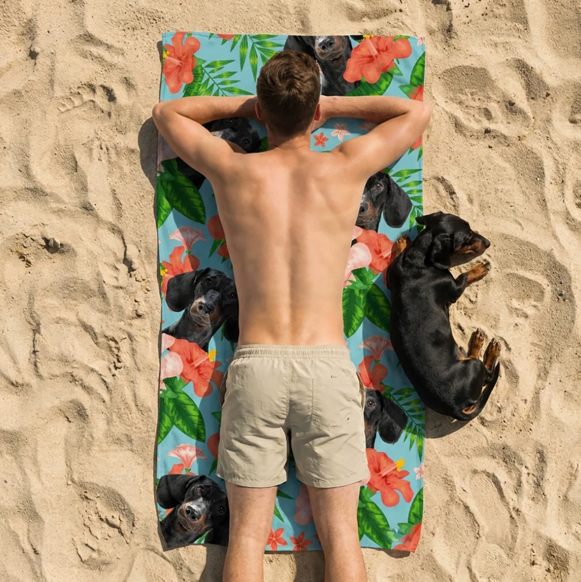 Persona tumbada sobre una toalla de playa personalizada con motivos de perros, rodeada de arena.