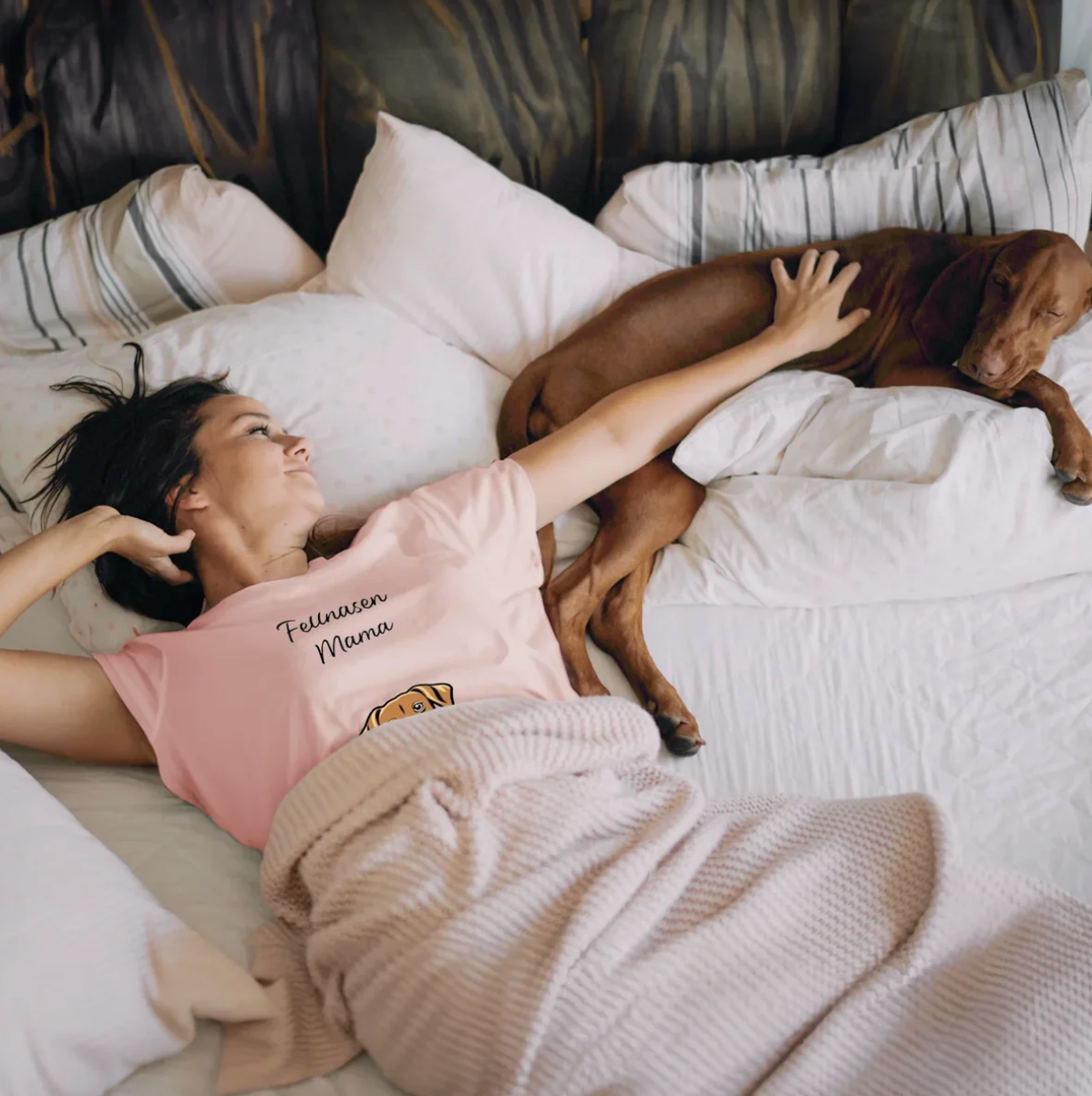 Mujer con camiseta rosa tumbada en la cama junto a un perro, rodeada de cojines y una manta.