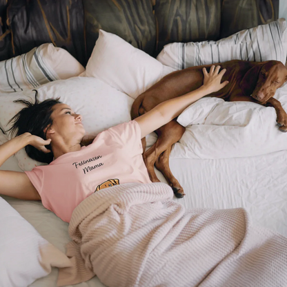 Mujer con camiseta rosa tumbada en la cama junto a un perro, rodeada de cojines y una manta.