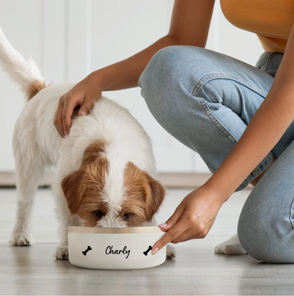 Perro comiendo de un comedero blanco personalizado con el nombre "Charly" y motivos de huesos.
